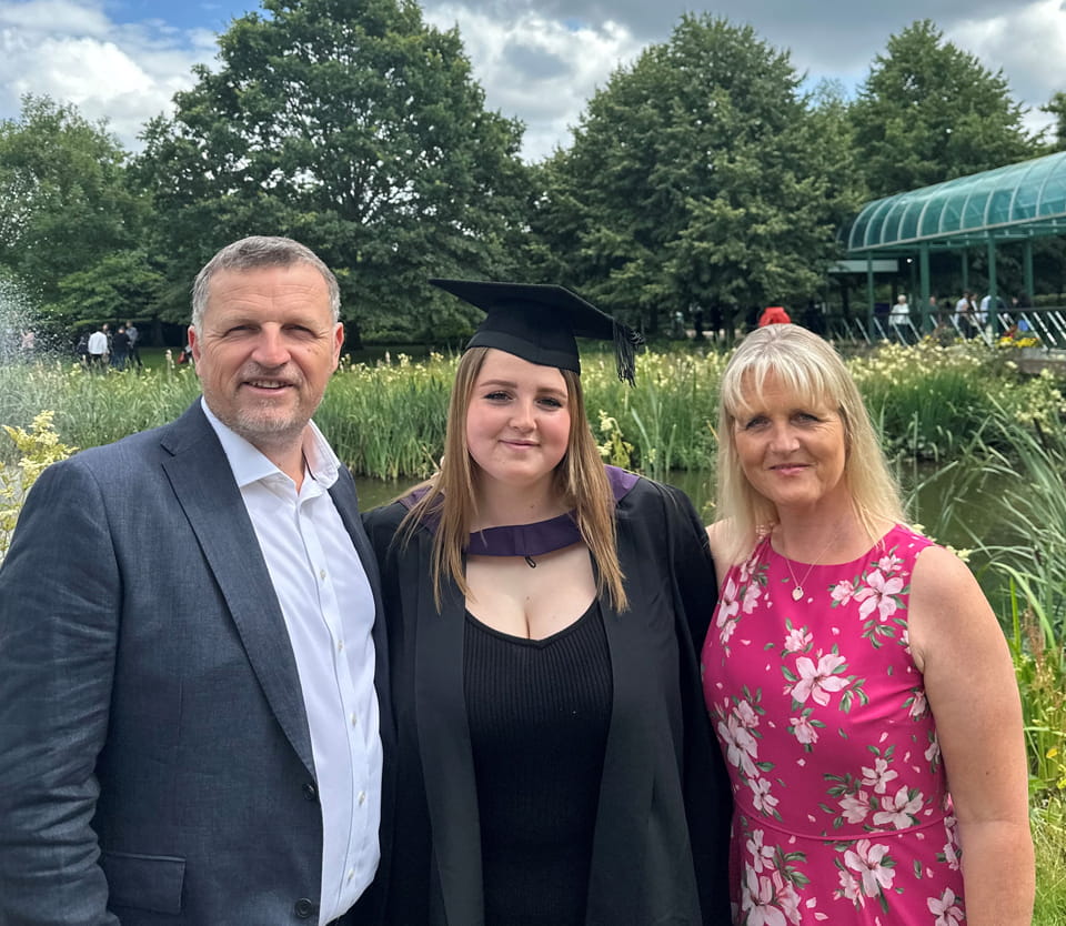 Chris, Molly and Sam Seal. Molly is wearing a graduation cap and gown. In the background there are trees, plants, and water. The photograph is taken at the west end of the campus, outside the Sir Denis Rooke building.